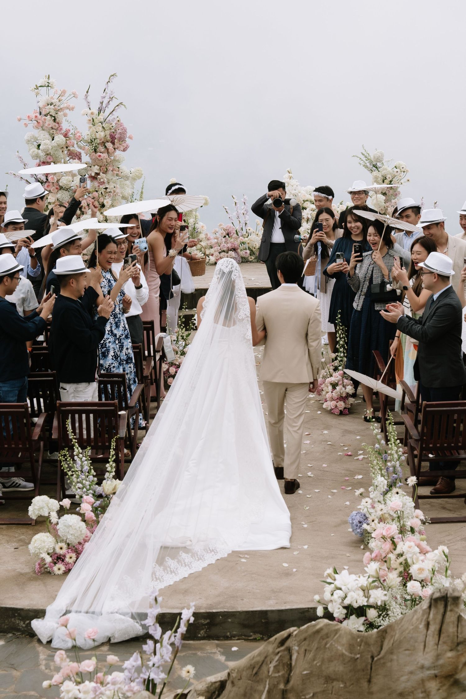Bride in a flowing veil walks down a floral-lined aisle toward the groom at an elegant outdoor mountain wedding ceremony.
