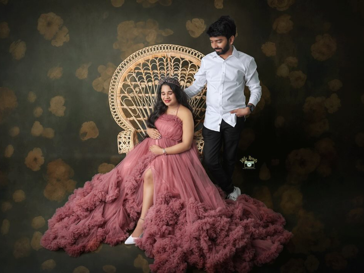 A romantic studio portrait with a decorative peacock chair and flowing pink gown against a brown backdrop.