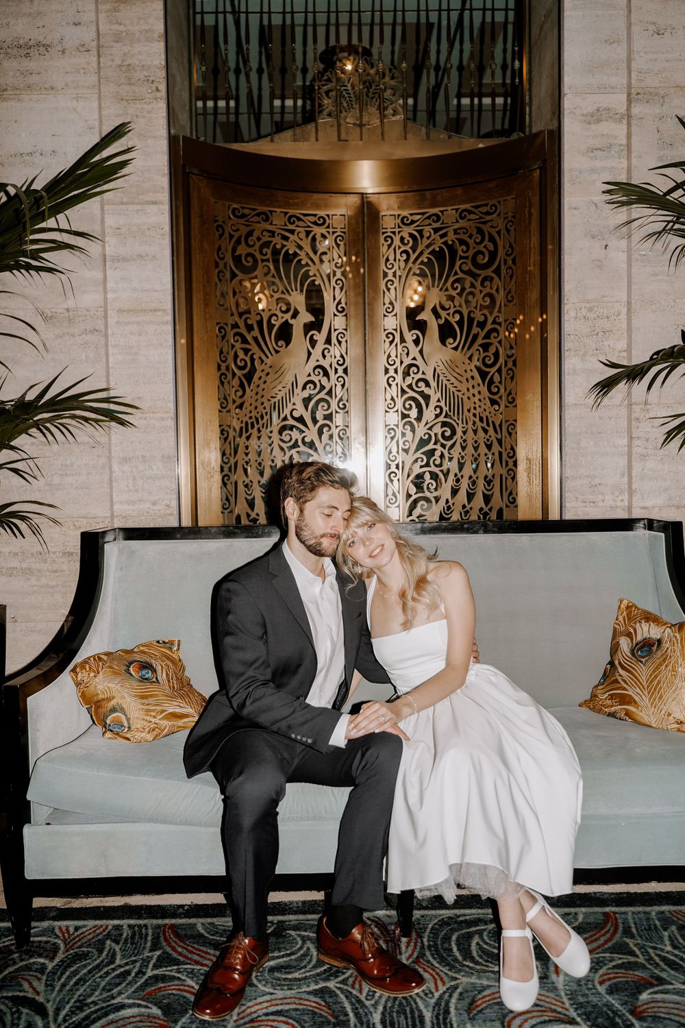 A couple in wedding attire shares romantic moments on an elegant blue sofa in front of ornate doors.