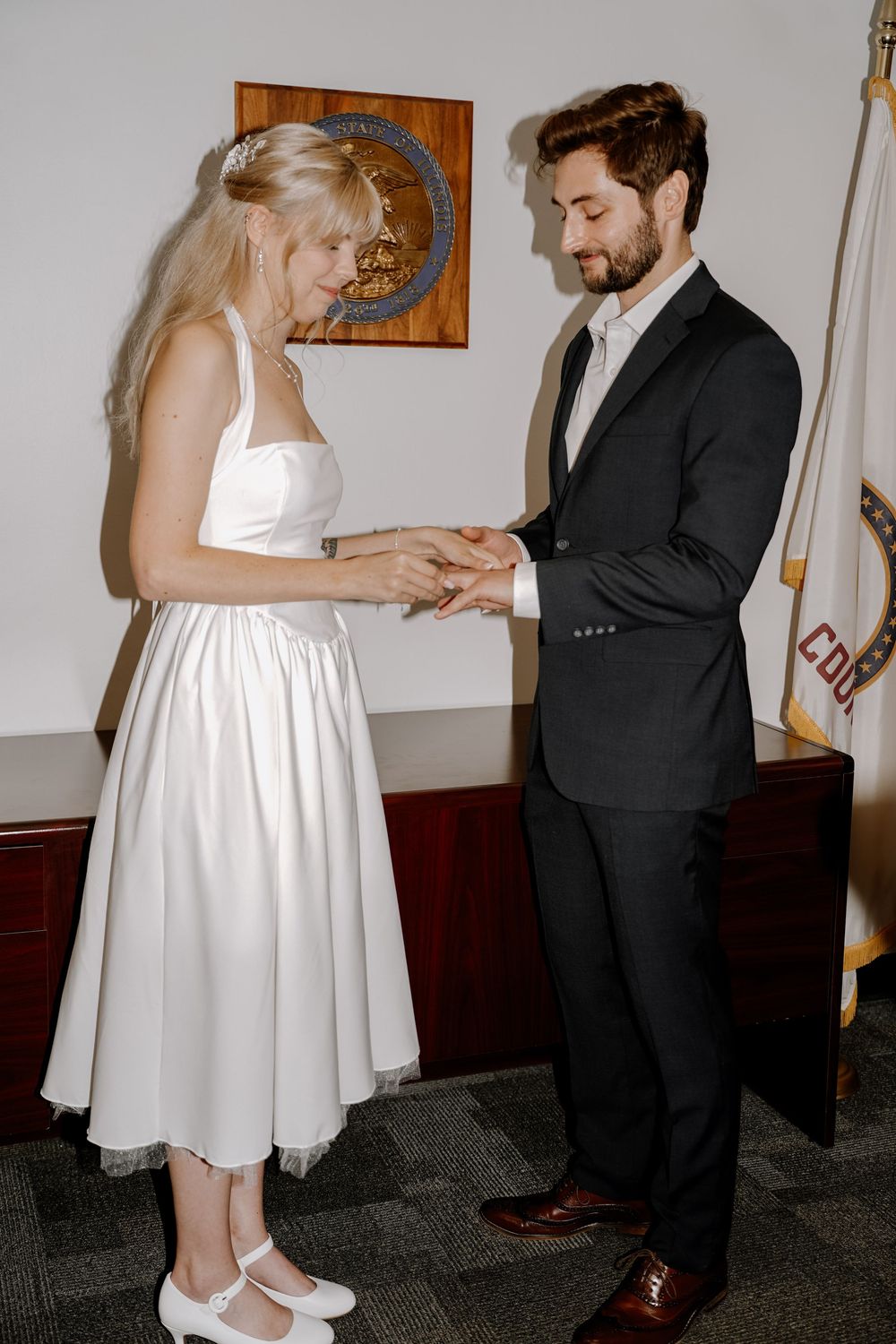 A couple in wedding attire exchanges rings during an intimate indoor ceremony with white dress and dark suit.