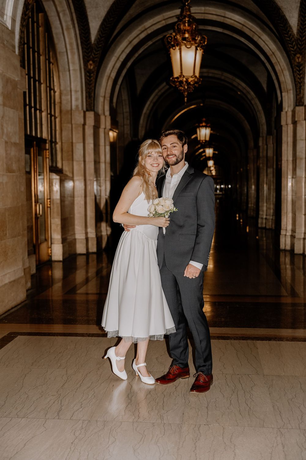 Couple shares romantic moments in elegant arched hallway with ornate lighting and marble floors during wedding celebration.