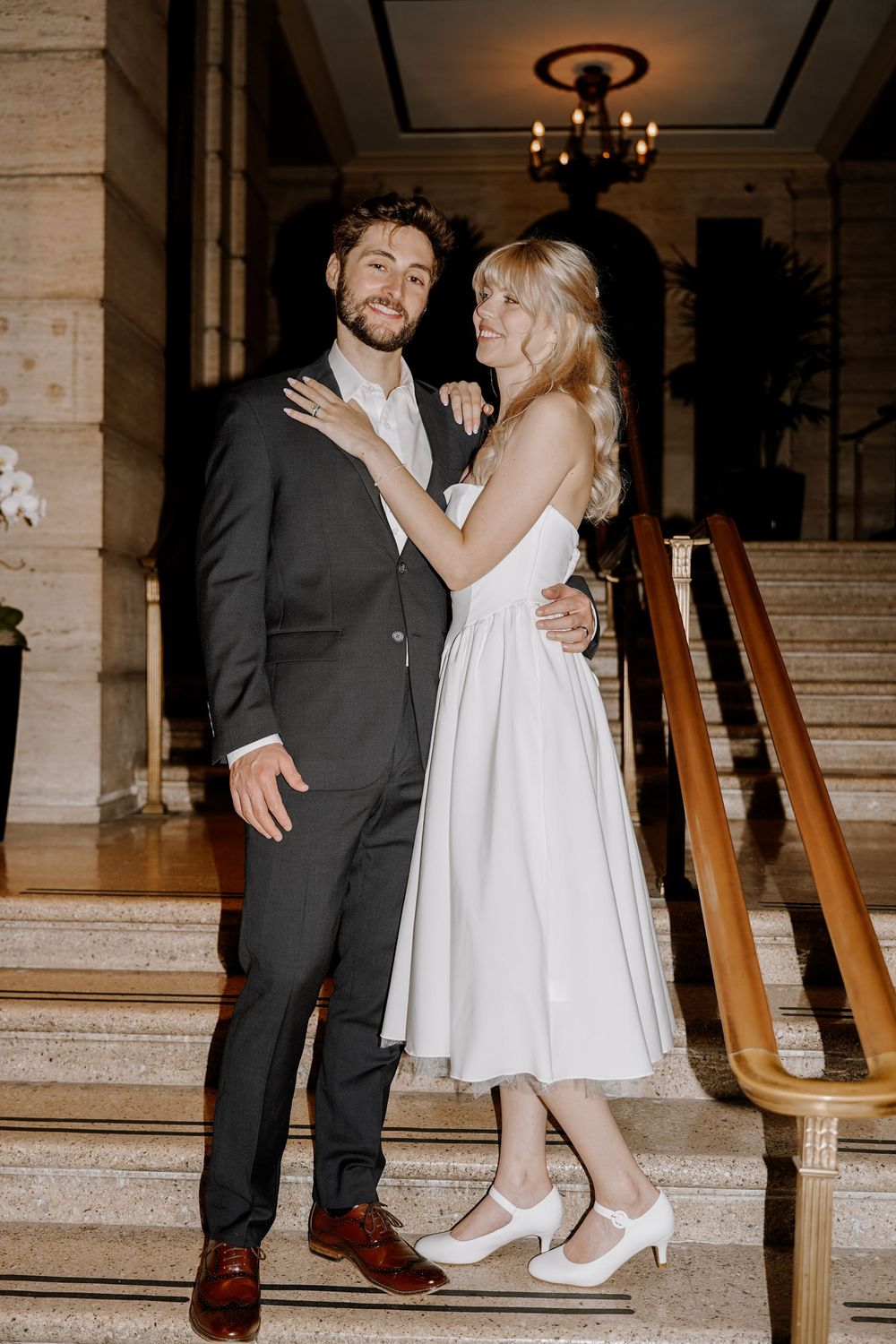 A couple in wedding attire dances together on marble steps in an elegant venue with a chandelier above.
