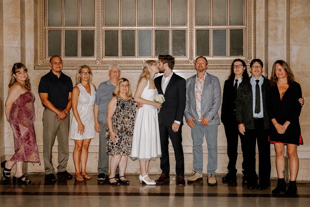 A group of formally dressed people stand in a line against a beige wall at an elegant indoor venue.