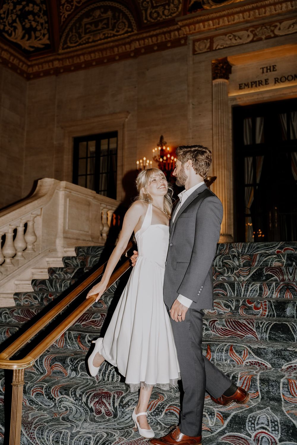 A couple in wedding attire dances on a grand staircase with ornate wrought iron railings in an elegant venue.