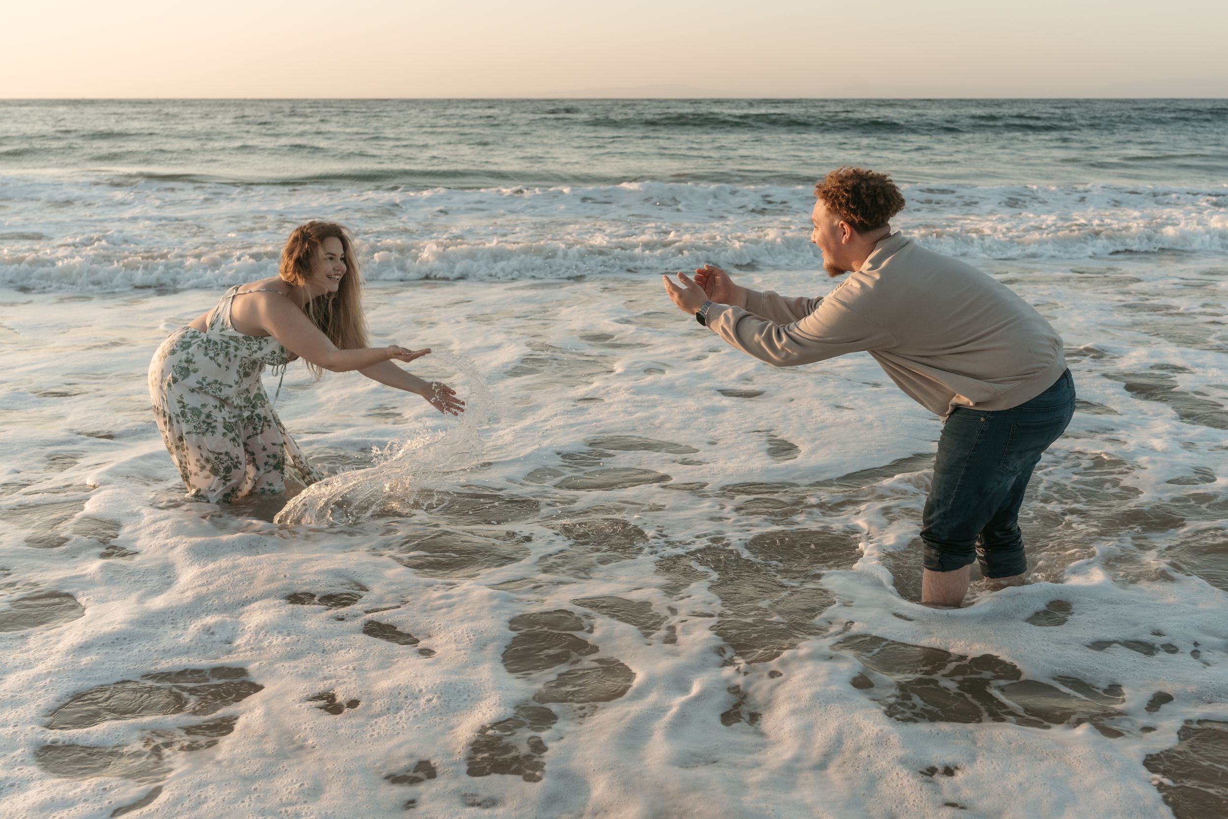 Two people joyfully playing in ocean waves on a sandy beach during sunset.