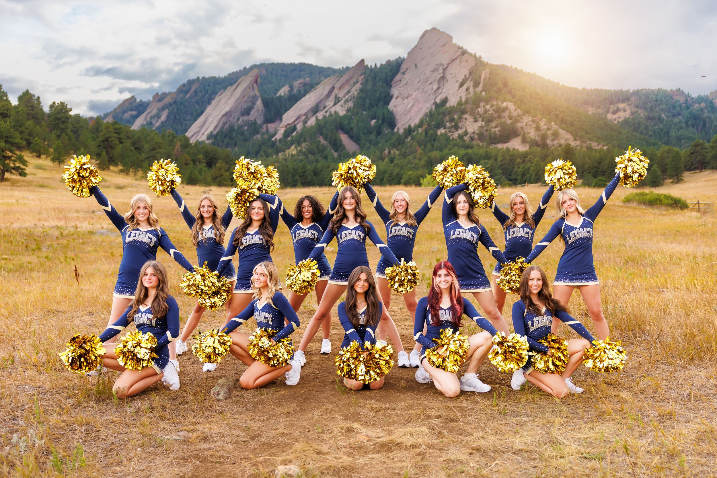 Cheerleaders in blue and gold uniforms pose with pom-poms in a field with the Flatirons mountains backdrop.