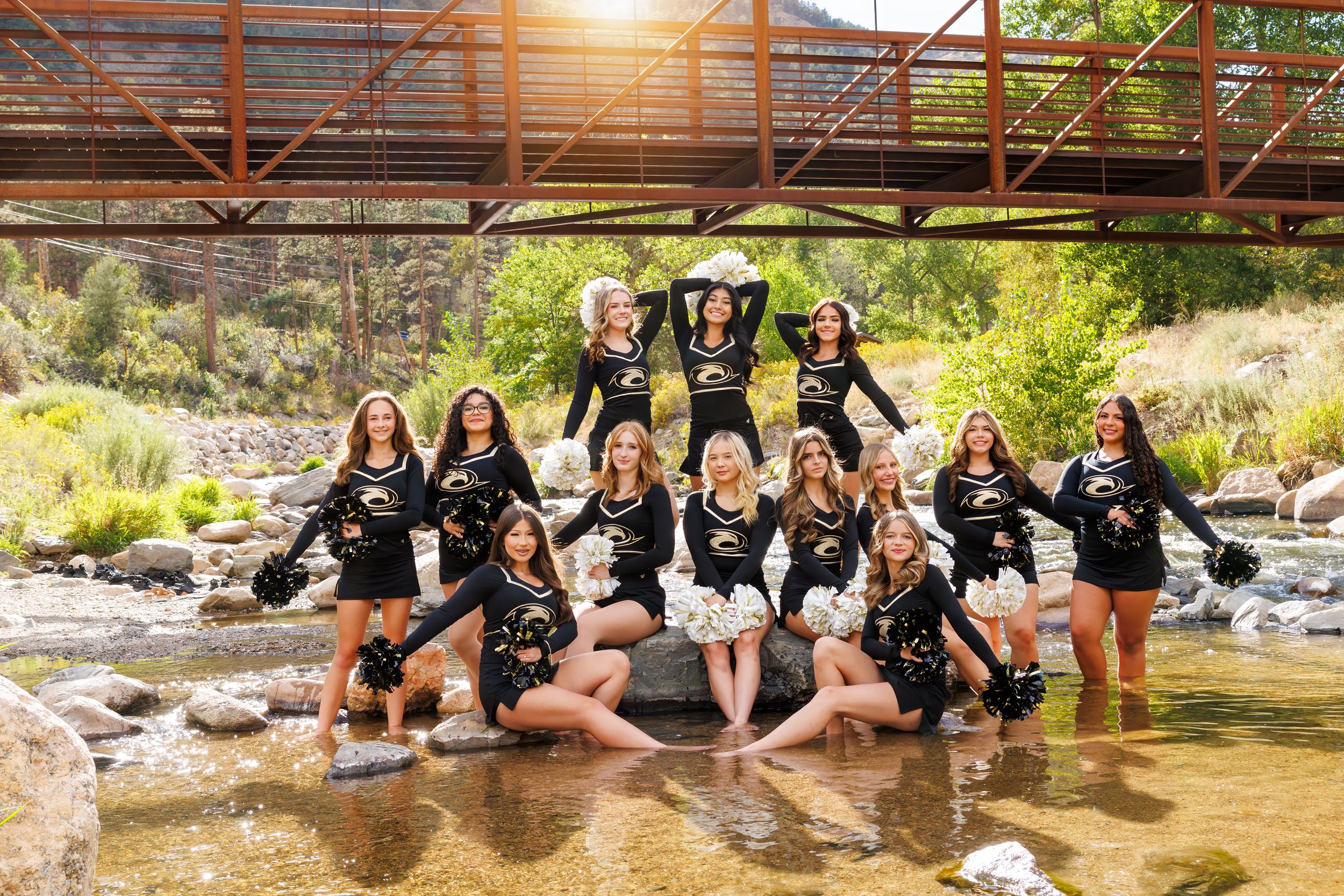 Cheerleading squad posing in black uniforms near a creek with a bridge backdrop outdoors.