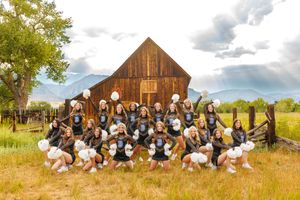 Cheerleading squad posing in uniform with pom-poms in front of rustic barn with mountain backdrop.