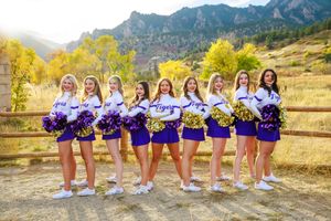A row of cheerleaders in purple and white uniforms pose together in front of scenic mountain landscapes.