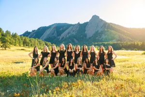 A row of people sitting in a meadow wearing black dresses against a mountainous backdrop at sunset.