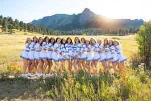 Large group wearing white dresses posed in row formation outdoors with mountains and sunlit trees in background.