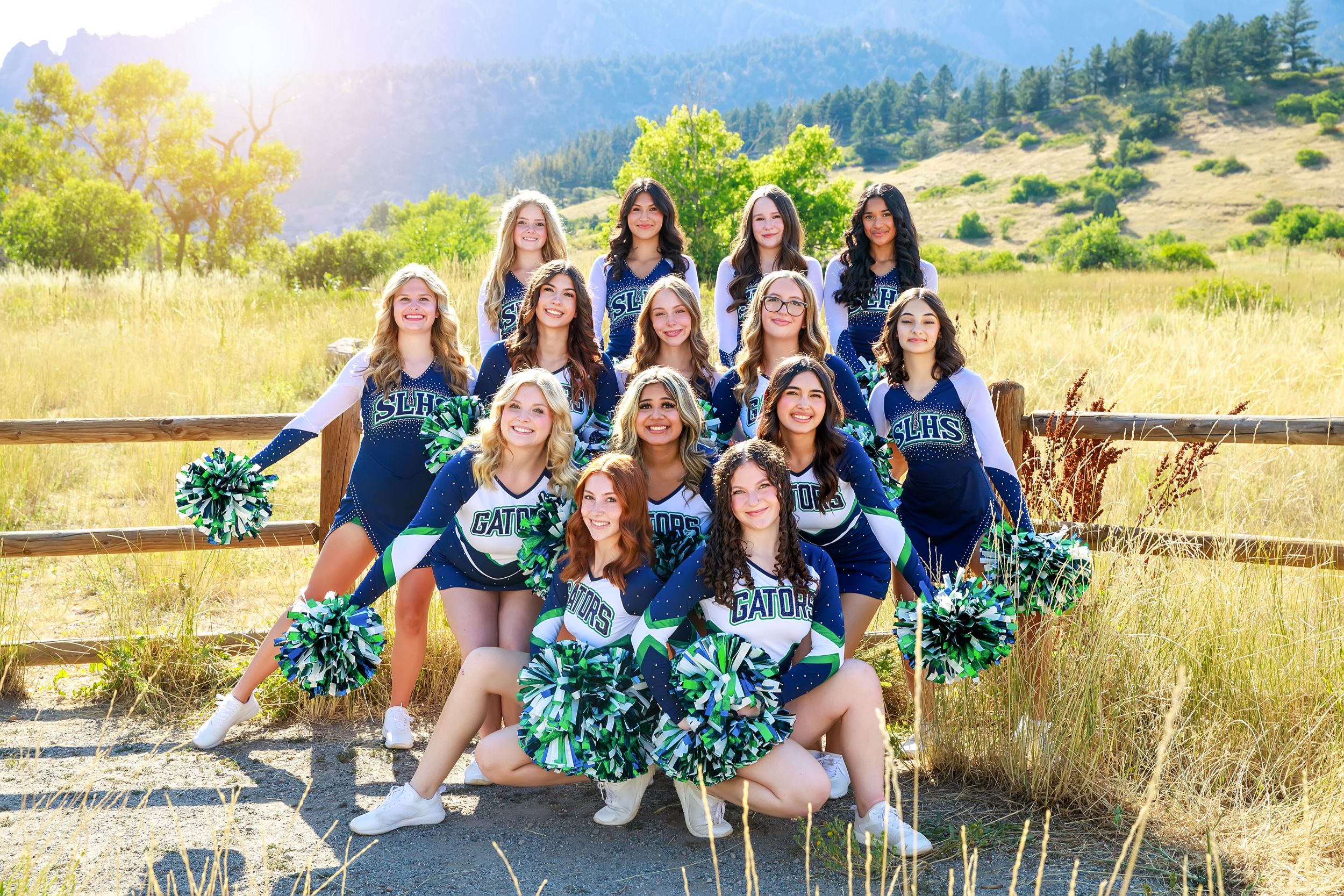 Cheerleaders in blue and white uniforms pose together outdoors against a scenic mountain landscape background.