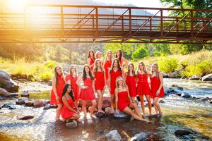 Group wearing red dresses poses together on rocks by flowing river during golden sunset hour.