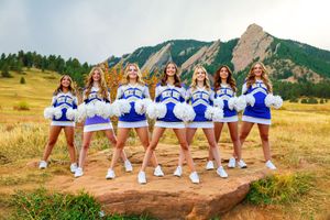 A group of cheerleaders in blue and white uniforms pose together against a scenic mountain backdrop at sunset.