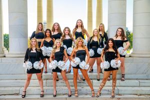 A dance team in black uniforms performs a high kick line choreography in front of white pillared building.