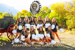 Native American themed dance team poses outdoors in traditional headdresses against autumn foliage backdrop.