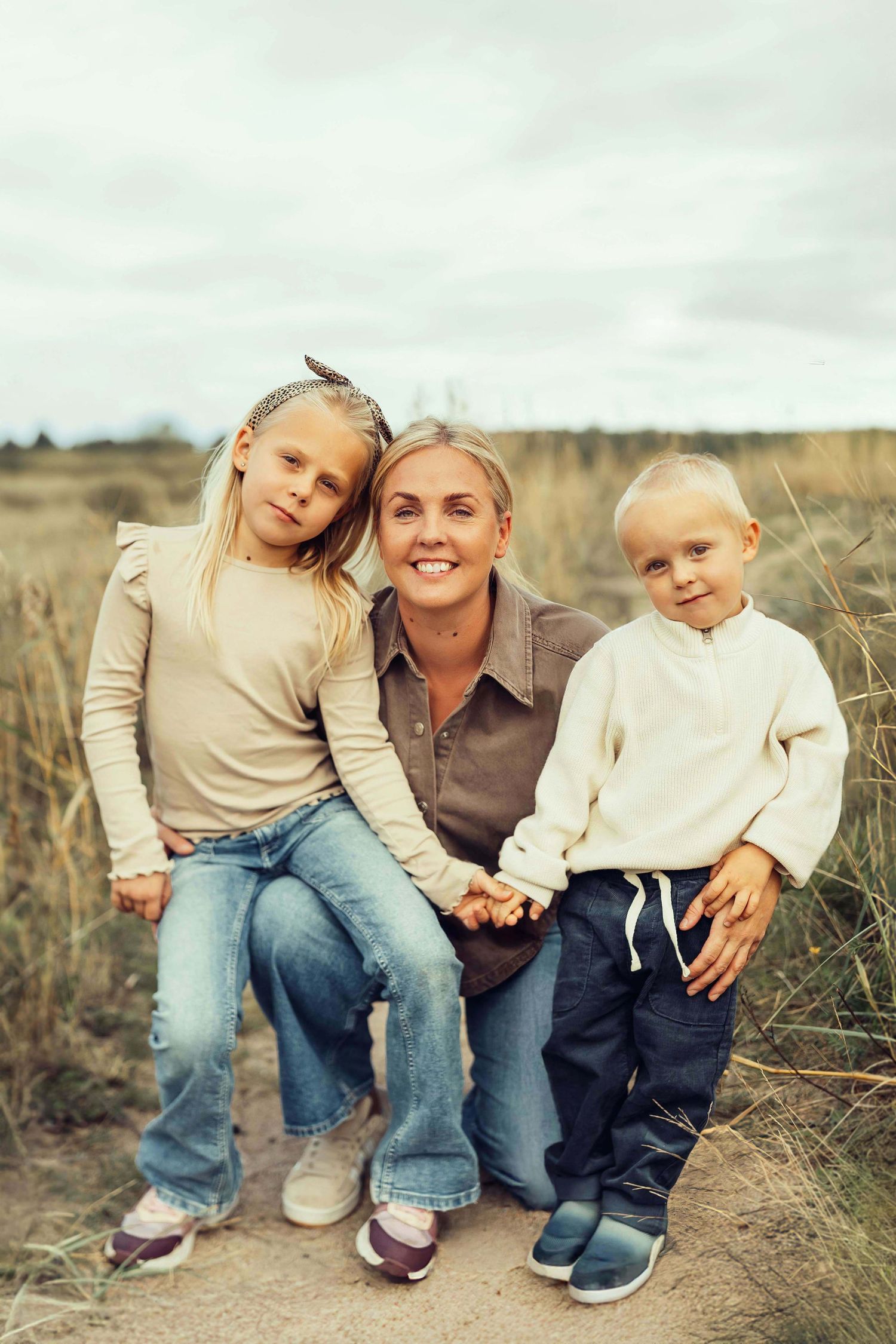 En leende mamma med sina två barn på varsitt knä, alla ler och tittar in i kameran