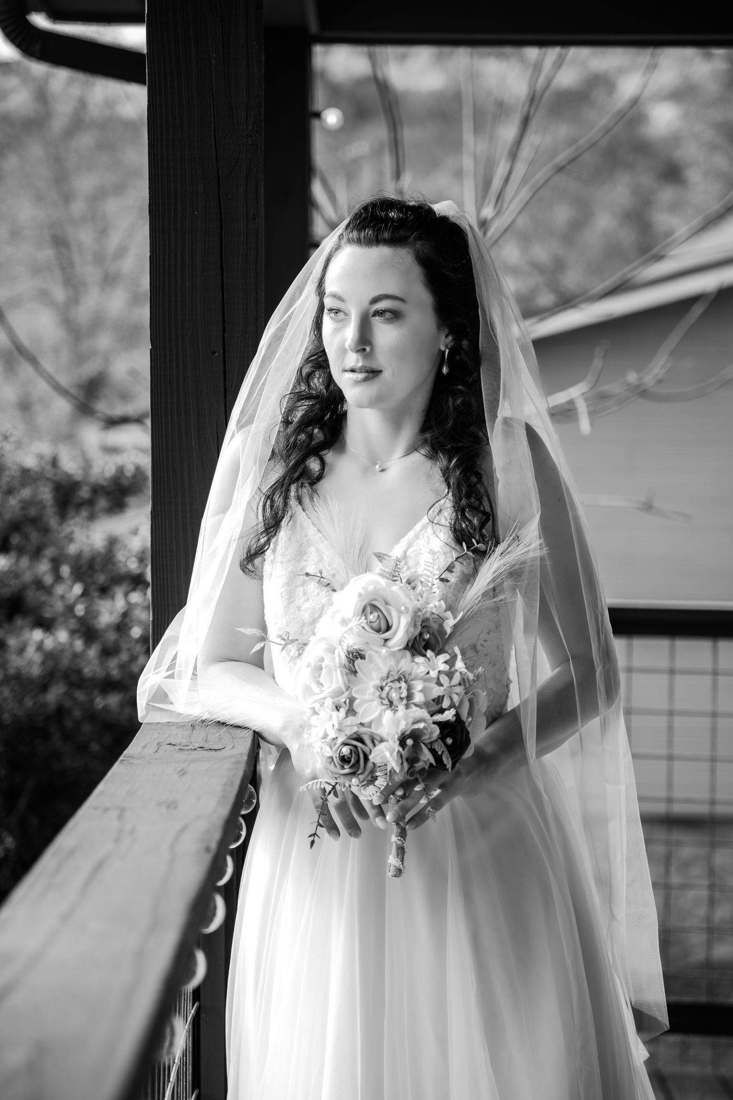 A bride in a flowing white gown and veil holds a rose bouquet while standing near a railing in black and white photography.