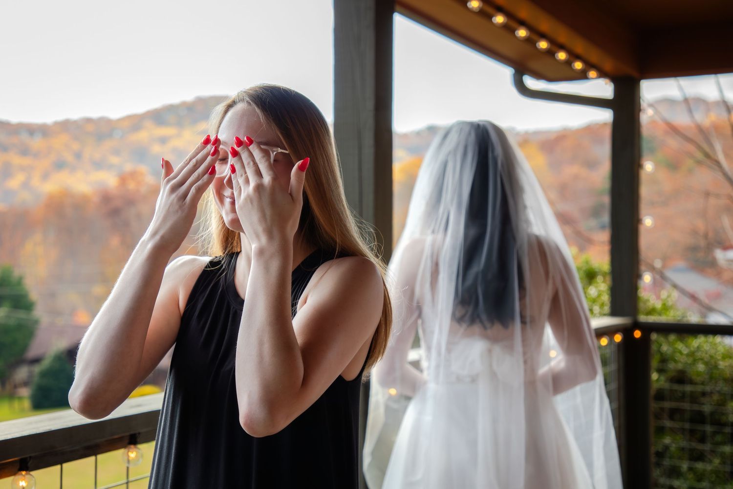 Bride in white wedding dress stands by window looking out at autumn foliage on a mountain landscape.