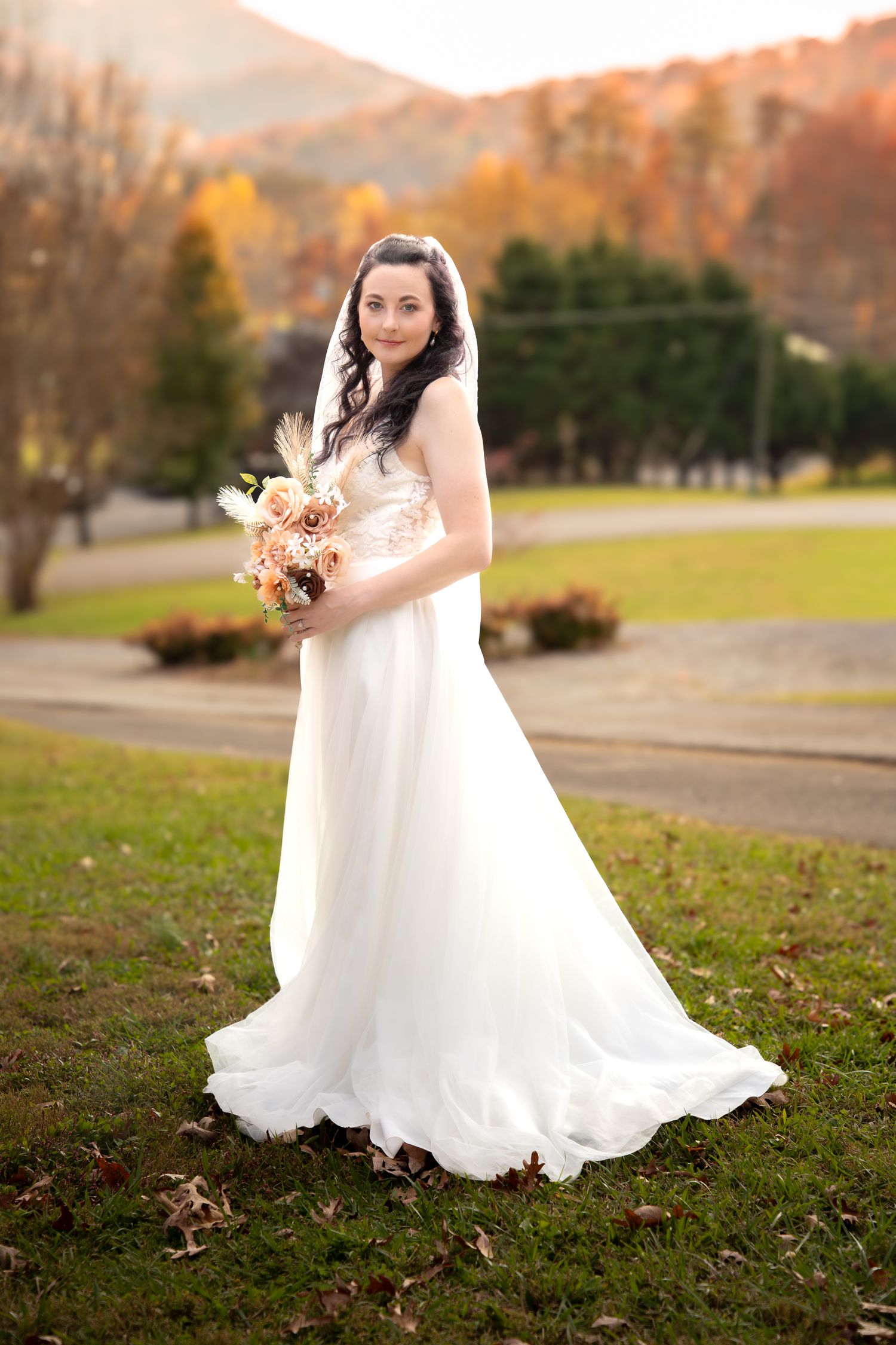 A bride in a flowing white wedding gown holds a dried flower bouquet while standing on grass during autumn.