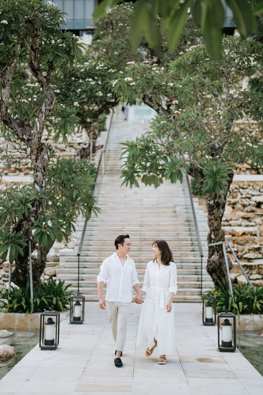 Couple in white outfits walking down garden steps decorated with lanterns.