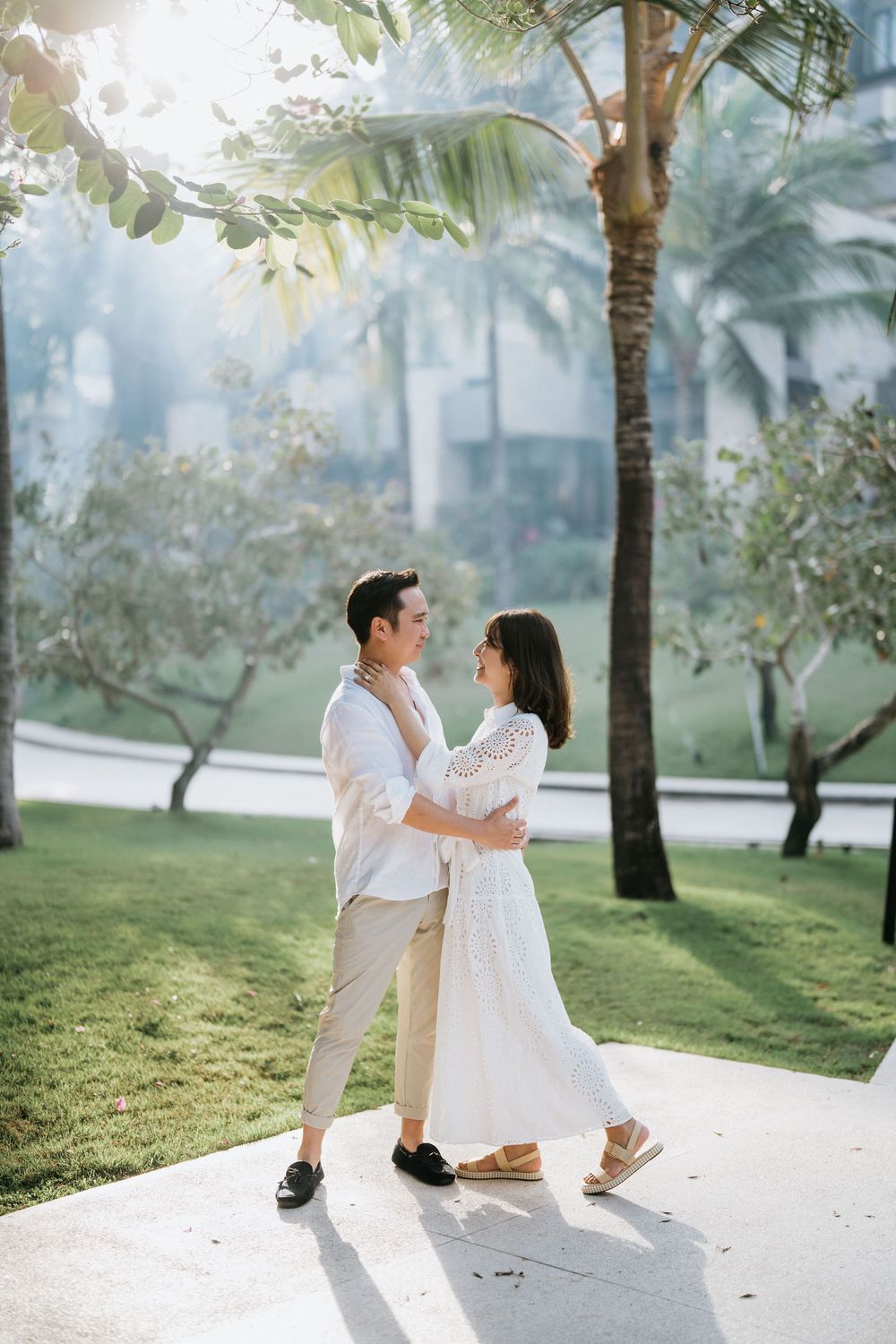 Romantic couple in white outfits embrace under palm trees in a tropical garden setting.