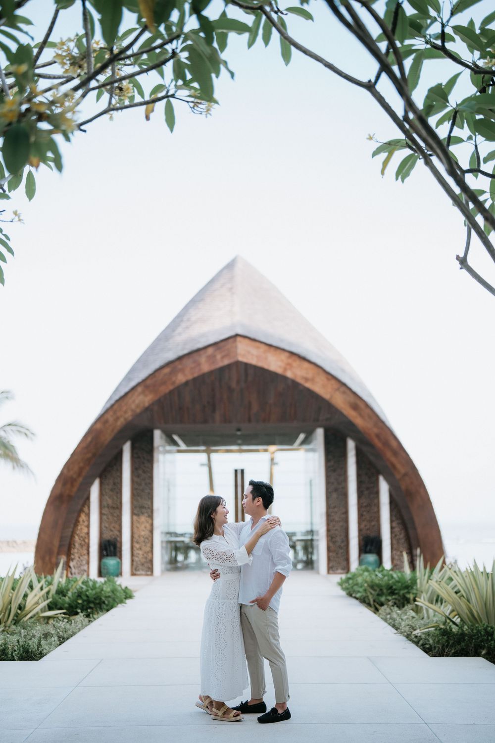 Couple embracing in front of an elegant arch-shaped chapel with tropical landscaping.