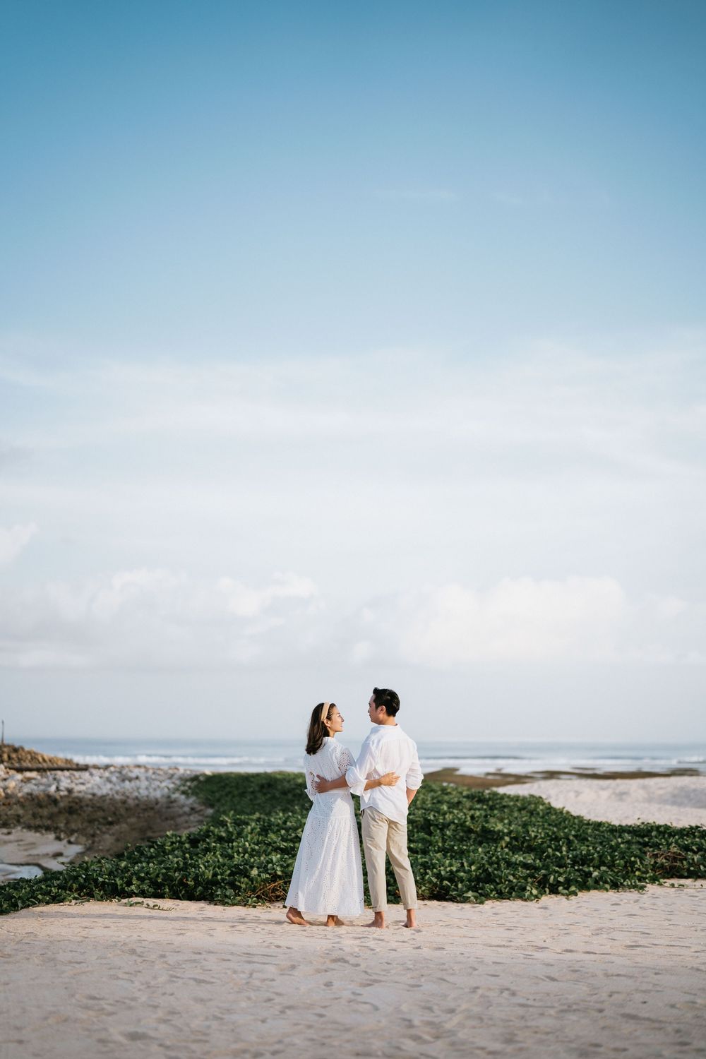Couple in white outfits sharing romantic moment on beach with ocean view.