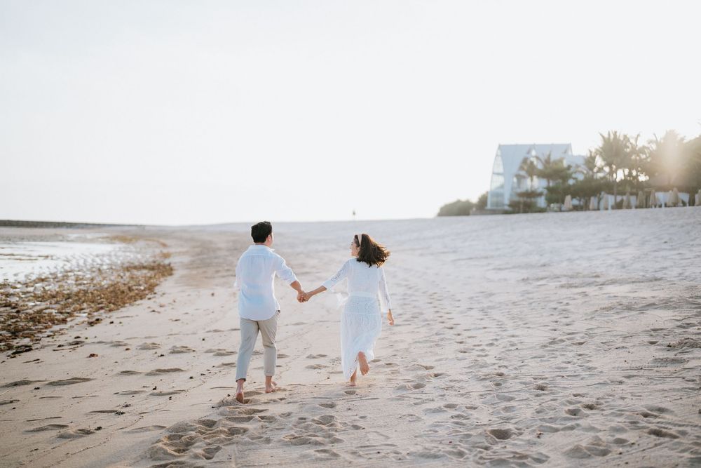 Couple in white and beige outfits walking playfully on a sandy beach at sunset.