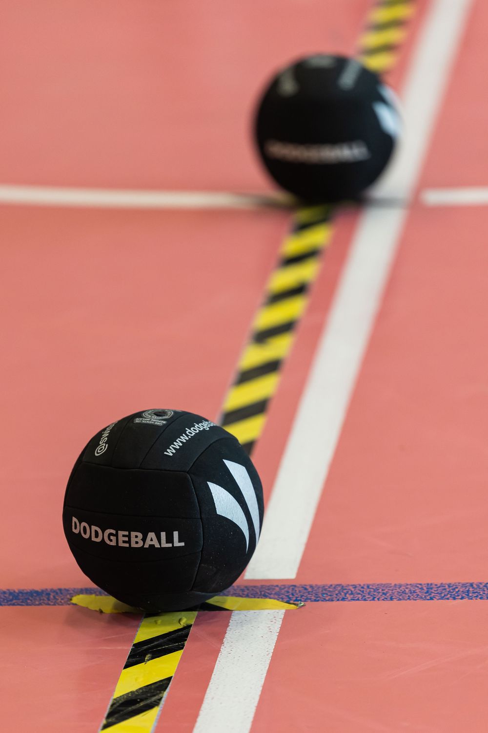 Black dodgeballs rest on a pink gymnasium floor with yellow and black striped caution lines and white boundary markings.