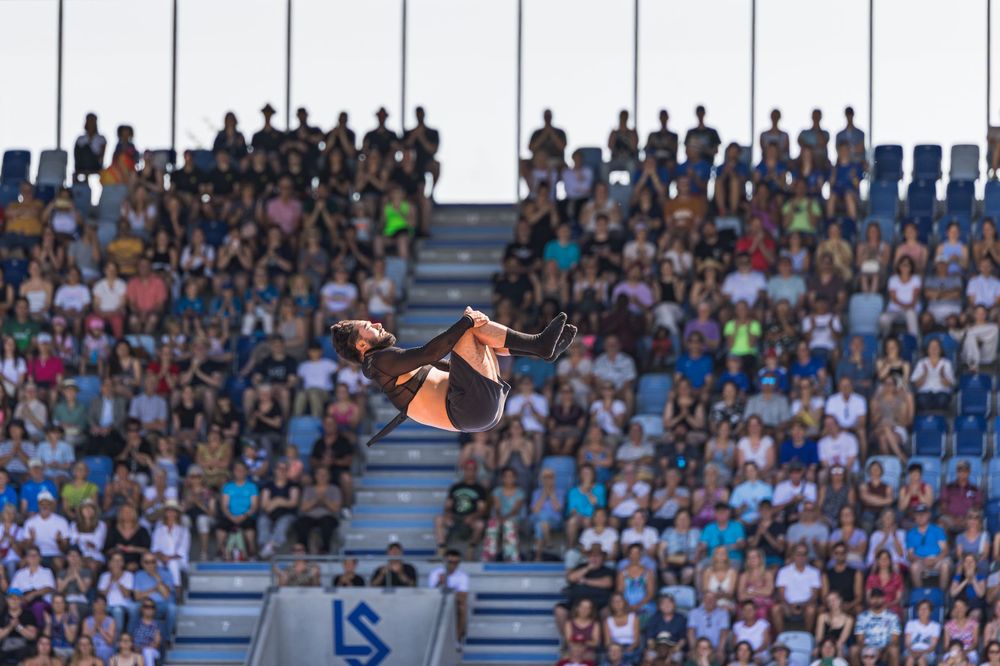 A gymnast soars through the air performing an acrobatic flip against a backdrop of stadium spectators.