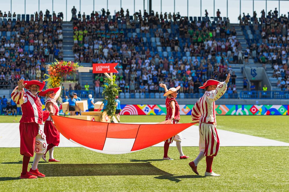 Performers in red and white striped costumes hold a large ceremonial banner on a sports field during an event.