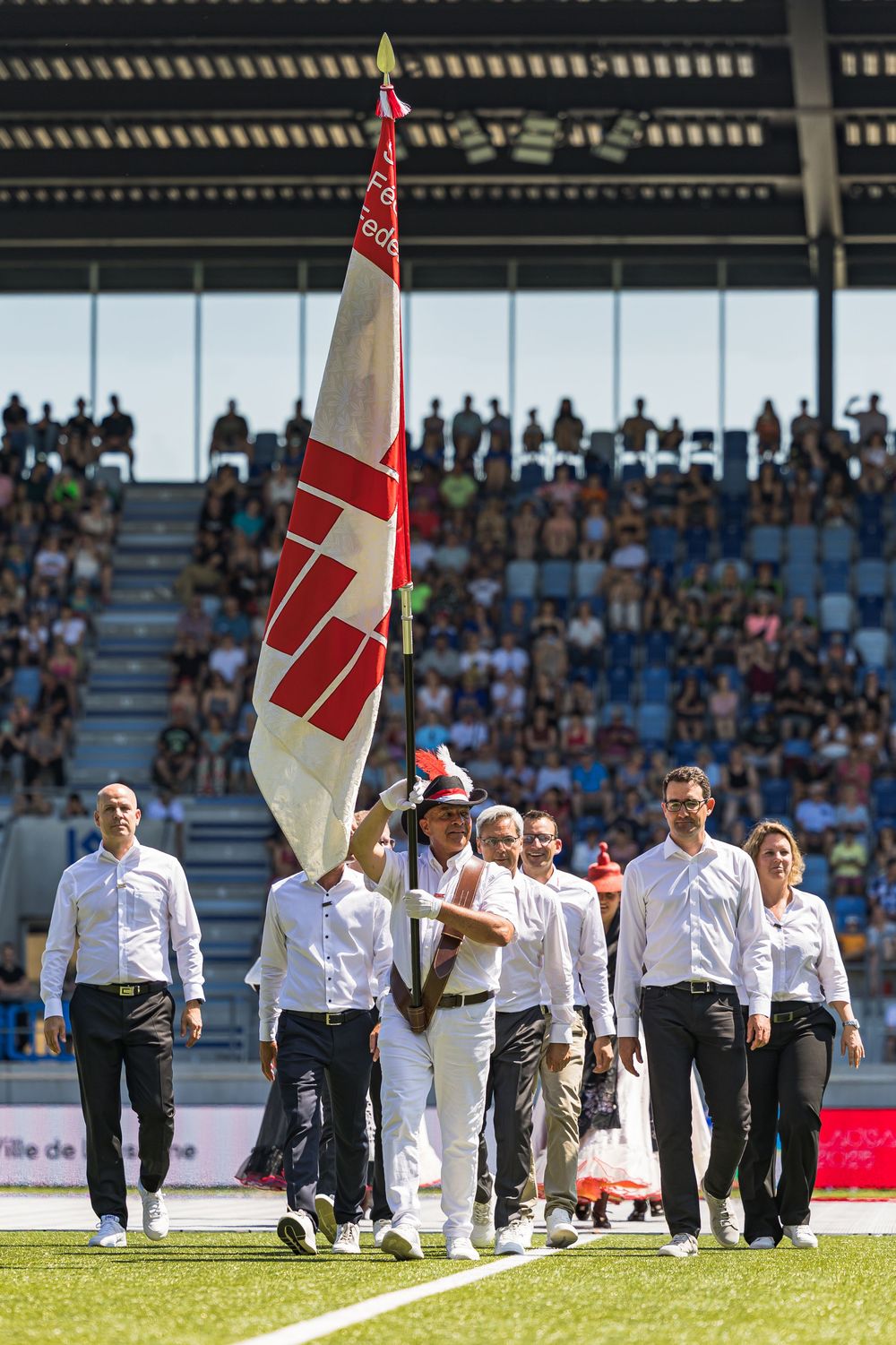 People walking in a line across a sports field carrying flags and banners during a sports ceremony.