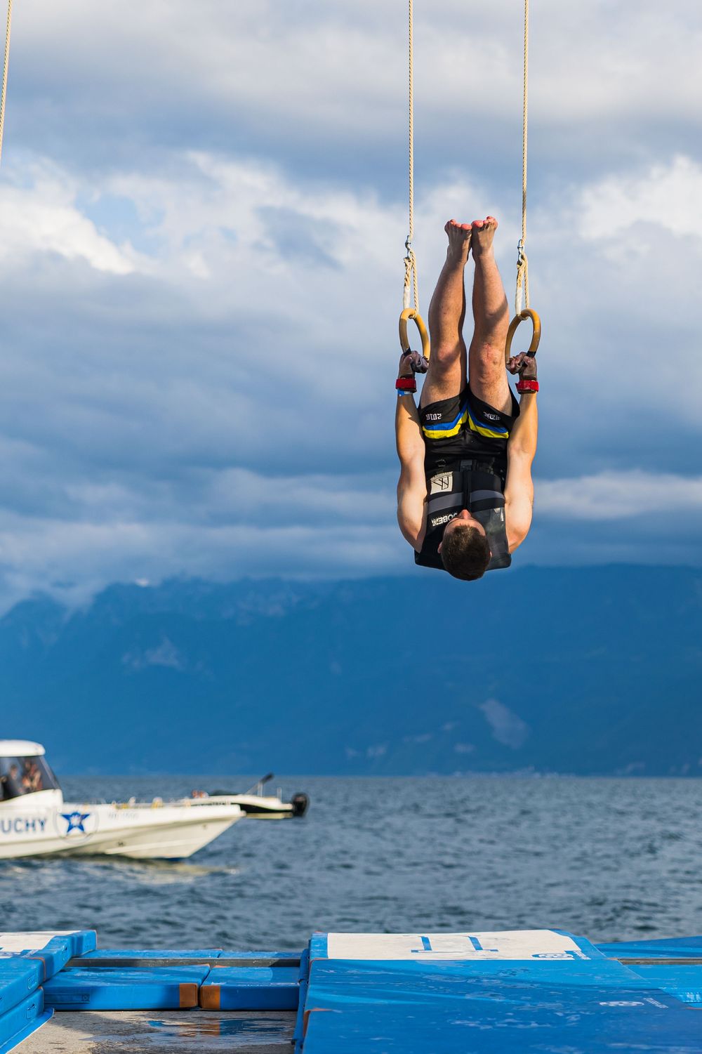 Gymnast performs on rings over ocean with boat and blue mats visible in background against dramatic cloudy sky.
