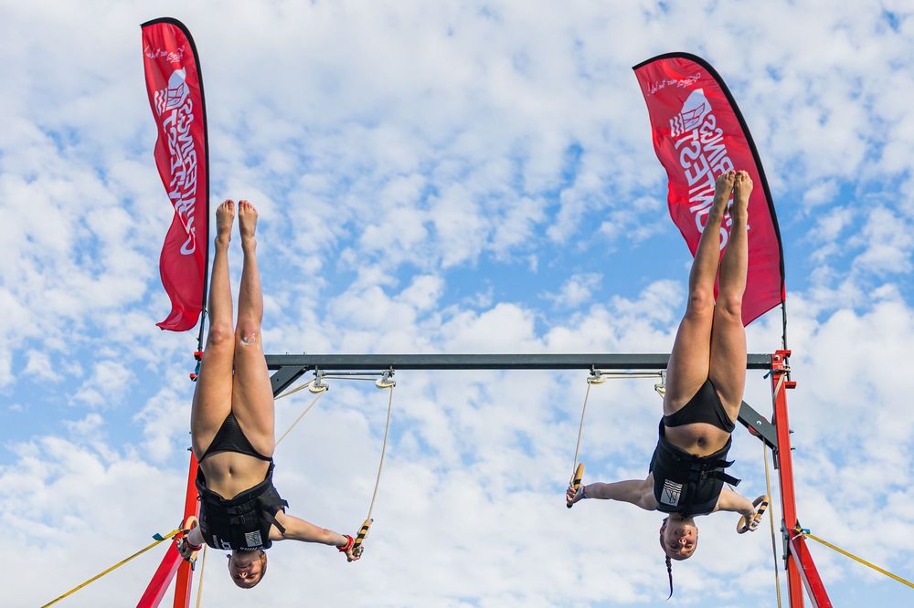 Two circus performers hanging upside down on aerial equipment with red banners against a cloudy blue sky.
