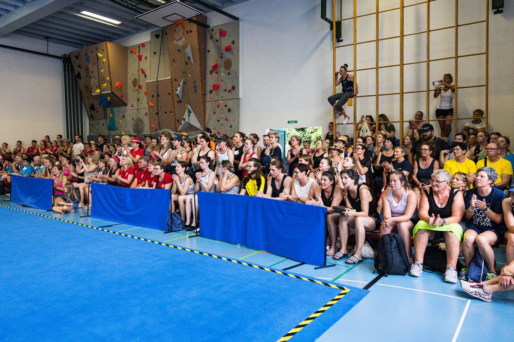 A large audience sits on mats watching climbers scale an indoor rock climbing wall with blue padding visible.