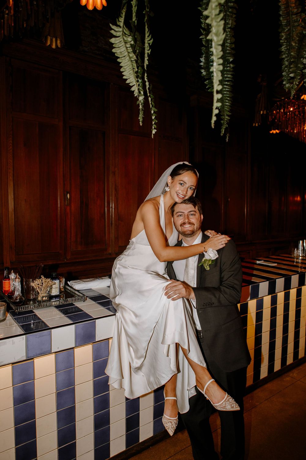 Playful wedding couple poses on striped banquette seating under hanging floral decor at reception venue.