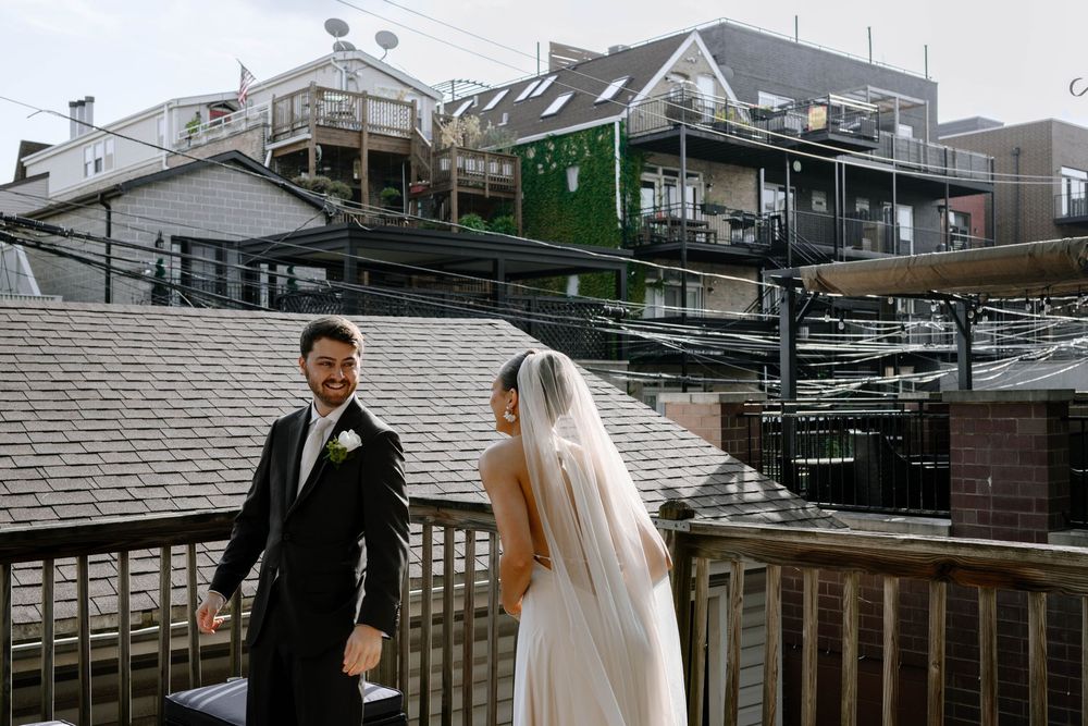 A couple in wedding attire share a moment on a wooden deck with multi-level buildings in the background.