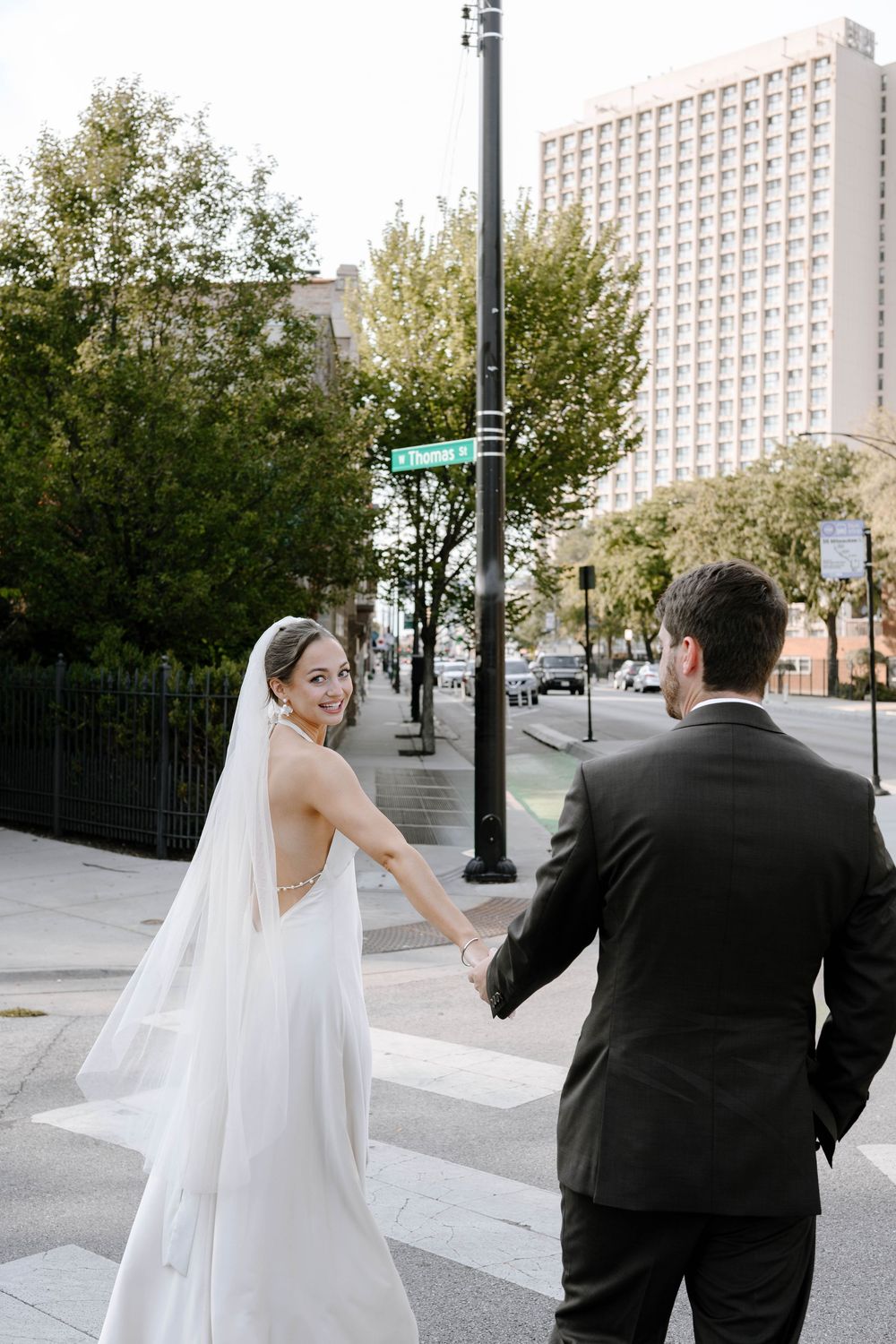 A couple in wedding attire walks down a city street with a tall building in the background.