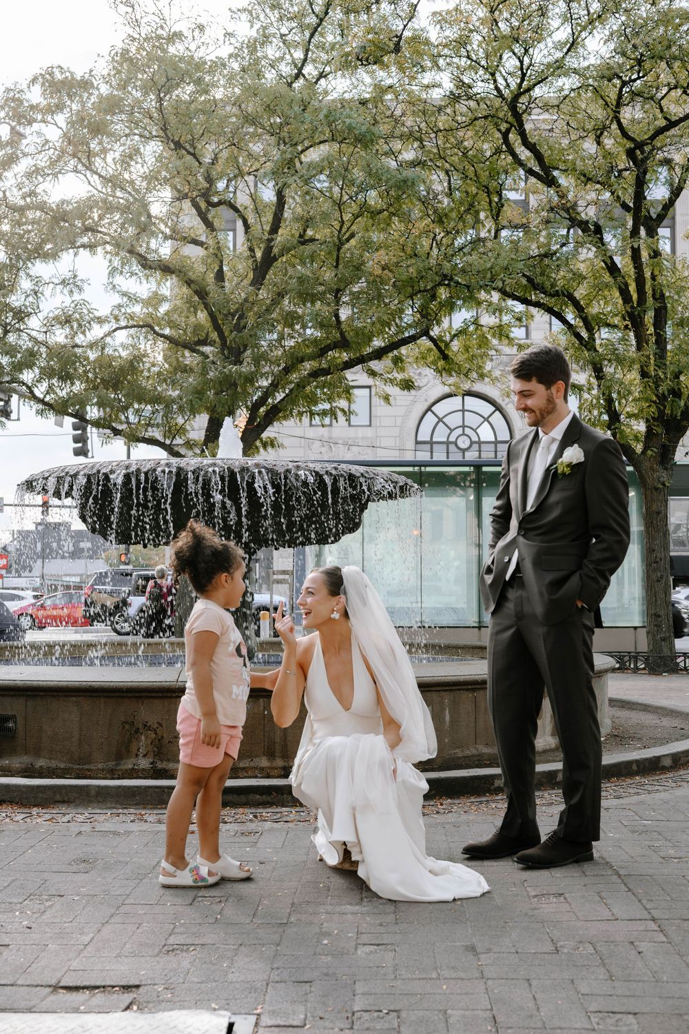 Wedding couple shares a sweet moment near an outdoor fountain with trees and guests in the background.