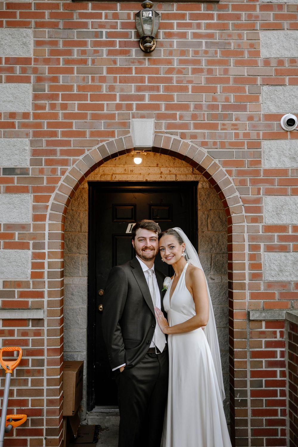 A couple poses together in front of a brick building with arched doorways during their wedding.