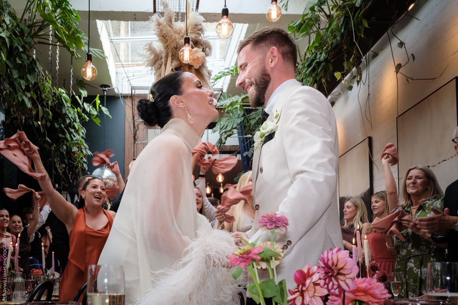 A joyful celebration scene at a wedding reception with guests dancing under bright pendant lights and lush greenery.