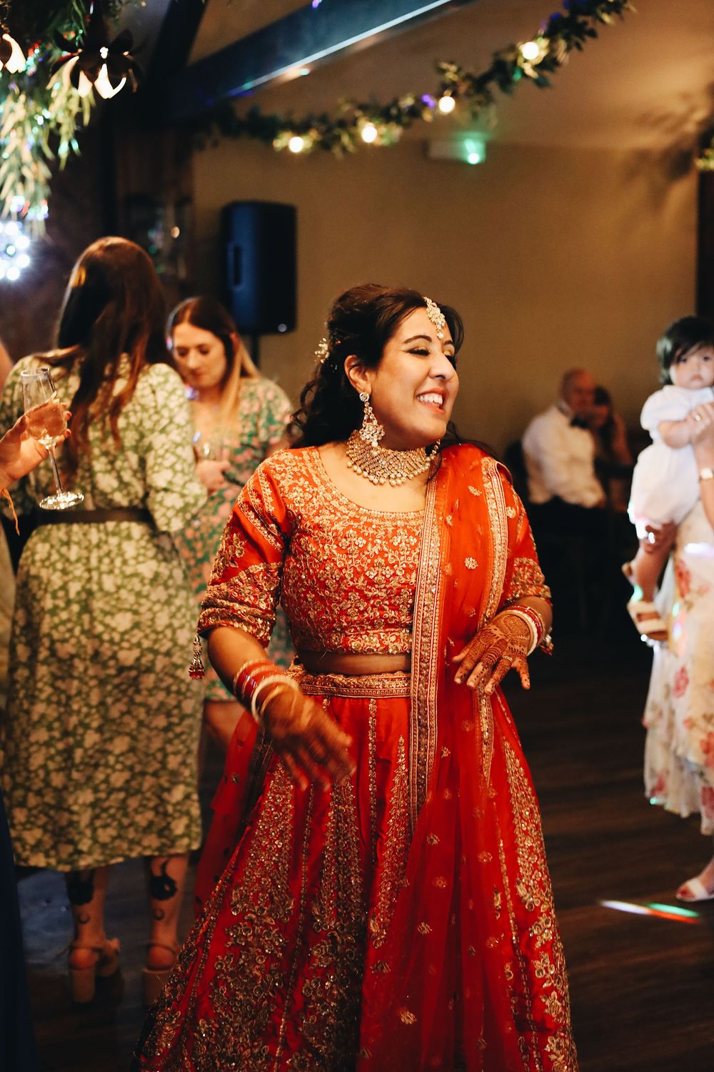 A guest dances joyfully at a wedding reception wearing a vibrant red and gold Indian sari with intricate embroidery.