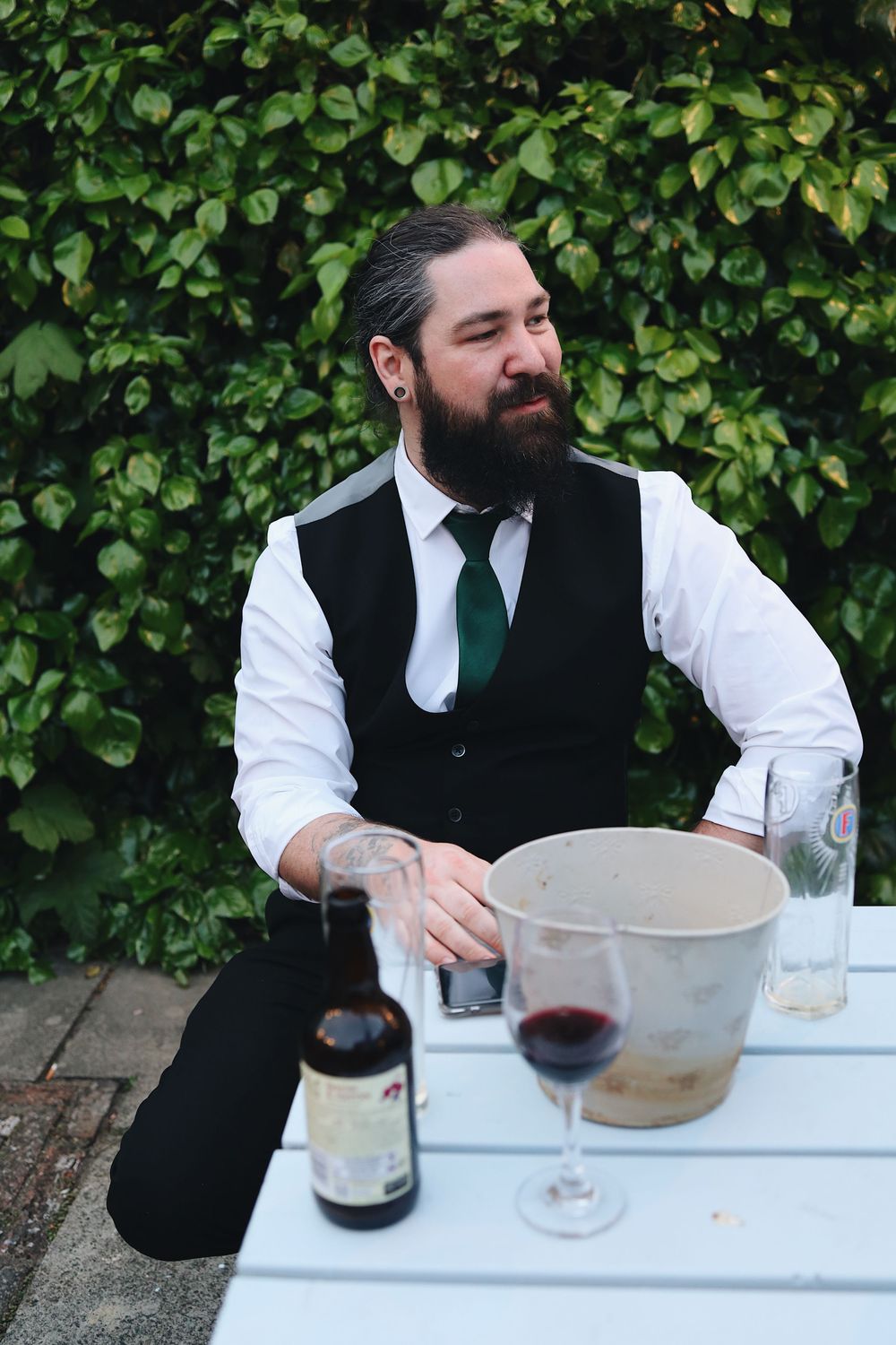 A person in formal attire sits at an outdoor table with wine glasses and beverages.