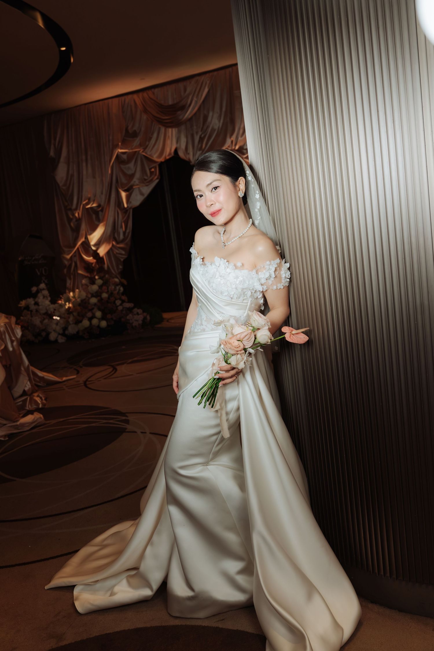Elegant bride in off-shoulder satin gown holds roses bouquet in warmly lit wedding venue.