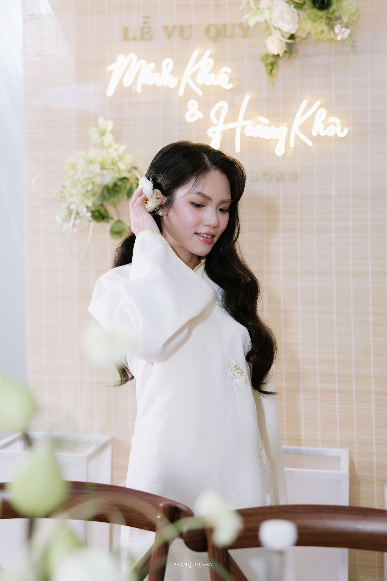 Elegant bride in white ao dai poses before neon sign reading Minh Khue and Hoang Kha at wedding.