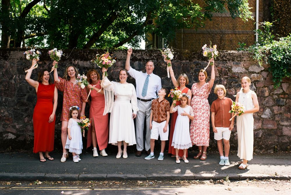 A cheerful group gathered outdoors raises their hands in celebration while standing against a brick wall on a sunny day.