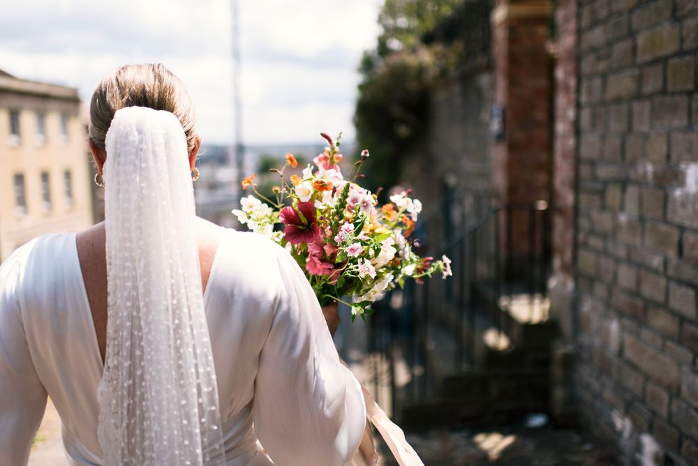 Bride holds a colorful bouquet in a white dress against a stone wall backdrop.