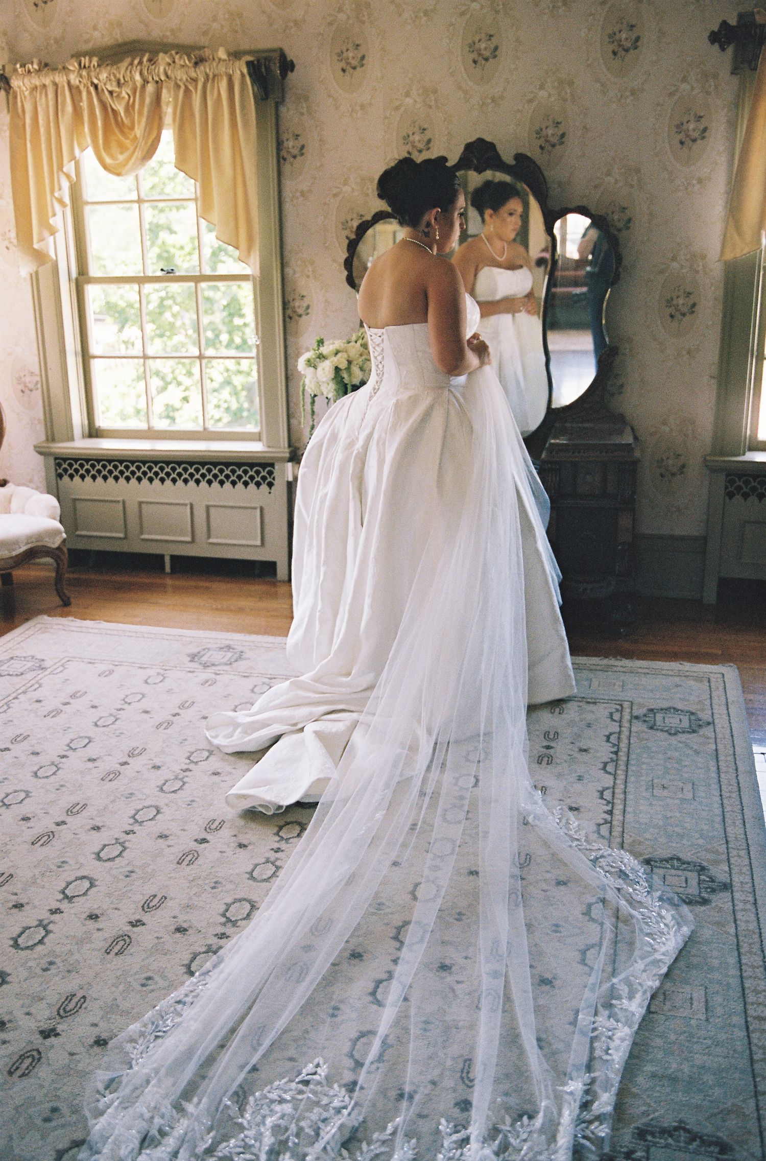 Bride in elegant white lace-trimmed wedding gown with long train, viewed from behind in a vintage mirror.