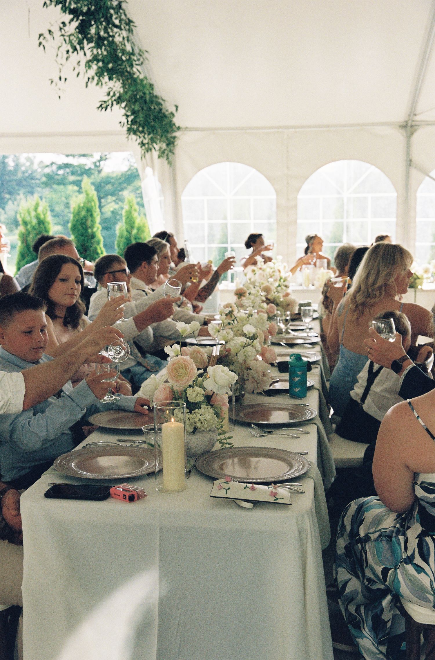 Guests raising glasses in a toast at an elegant wedding reception under a white tent with floral centerpieces.